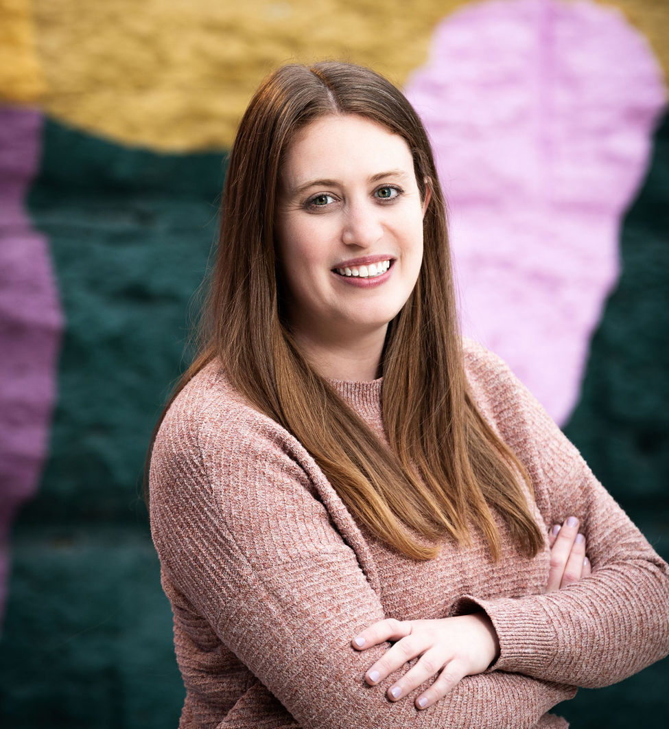 Susan stands with her body angled away in front of a mural with her arms crossed, smiling directly at the camera