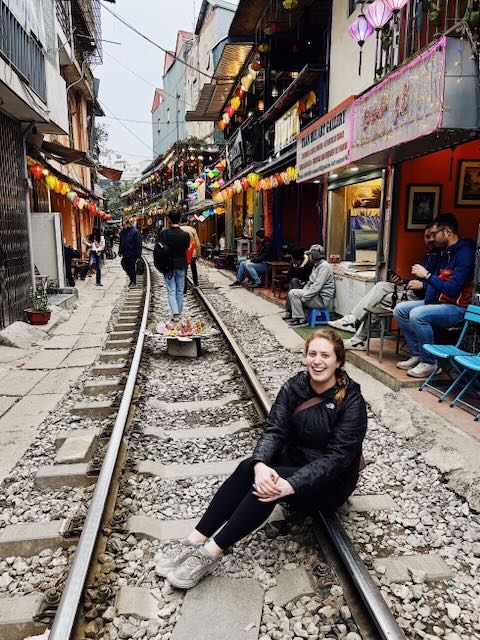 Susan sits on one side of a pair of railroad tracks in Hanoi, Vietnam, surrounded by small cafes on both sides.