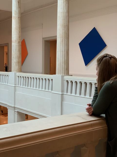 Susan faces away from the camera and leans against a half-height wall at the Art Institute of Chicago. A colonade with large, single-color pieces of art is visible beyond.